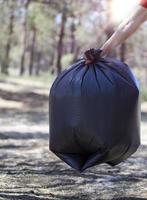 hand innehar mot en skog full av sopor en stor svart plast väska foto