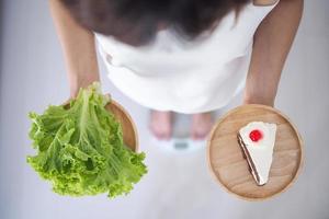 kvinna innehav vegetabiliska maträtter och kakor tänkande av välja till äta som är Mer välgörande till de kropp. vikt förlust begrepp, äter saker den där göra friska foto