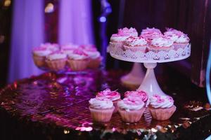 uppsättning av olika läckra läckra muffins på bord festlig bakgrund med glänsande rosa duk. olika desserttarteletter med dekorerad grädde och rosa kulisser. selektiv fokusering. godisbar koncept foto