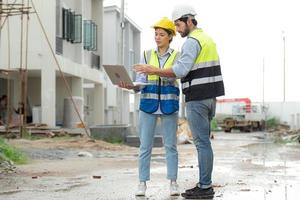 ingenjör man och kvinna arkitekt ha på sig säkerhet hjälmar diskutera hus utveckling projekt på konstruktion webbplats använder sig av bärbar dator dator. entreprenör chef granskning byggnad egendom infrastruktur. foto