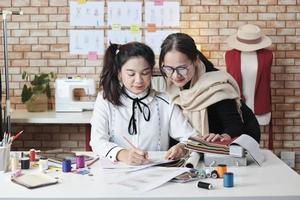 asiatisk medelålders kvinna mode designer lär en ung tonåring praktikant skräddare i studio med färgrik tråd och sömnad tyg för klänning design samling idéer, professionell boutique små företag. foto