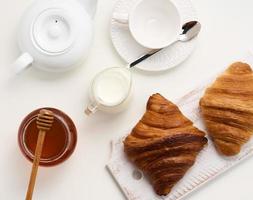 bakad croissanter, vit keramisk tekanna och tömma kopp och fat, burk av honung på en vit tabell foto