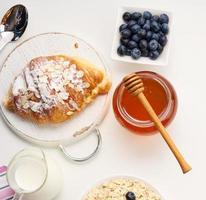 morgon- frukost, rå gröt flingor i en keramisk tallrik, mjölk i en karaff, blåbär och honung i en burk på en vit tabell foto