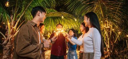 porträtt av Lycklig asiatisk grupp av vänner har roligt med sparklers utomhus- - ung människor har roligt med fyrverkeri på natt tid - människor, mat, dryck livsstil, ny år firande begrepp. foto