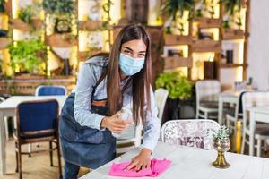 servitris bär skyddande ansikte mask medan desinficerande tabeller på restaurang eller caffee för Nästa kund. korona virus och små företag är öppen för arbete begrepp. foto