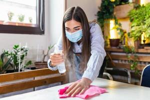 servitris bär skyddande ansikte mask medan desinficerande tabeller på restaurang eller caffee för Nästa kund. korona virus och små företag är öppen för arbete begrepp. foto