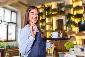 ung kvinna arbetssätt innehav en bricka med kaffe och glas av vatten. skön servitris bär förkläde och vinka.porträtt av en leende servitris innehav bricka av drycker i restaurang foto