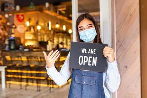 Lycklig kvinna servitris med skyddande ansikte mask innehav öppen tecken medan stående på Kafé eller restaurang dörröppning, öppen om igen efter låsa ner på grund av till utbrott av coronavirus covid-19 foto