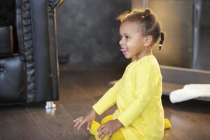 liten afrikansk amerikan flicka i en gul klänning med lockigt pigtails visar henne tunga. foto