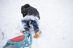 barn i vinter. de pojke är pulka. en barn täckt i snö klättrar en berg och drar en kälke Bakom honom. foto