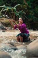asiatisk kvinna spelar med smutsig vatten från en smutsig flod medan bär en lila klänning och grön kjol foto