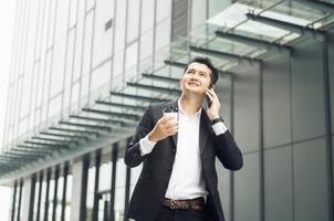 stilig leende asiatisk affärsman innehav smartphone i kontor affärsman ser på hand innehav kaffe kopp medan stående ensam i modern arbetsplats. foto