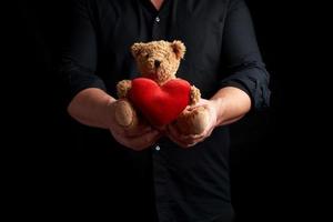vuxen man i en svart skjorta innehar en brun teddy Björn med en röd hjärta på en mörk bakgrund foto