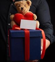 man i en svart skjorta innehar en blå fyrkant låda bunden med en röd band och sitter på topp av en brun teddy Björn med en hjärta foto