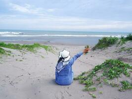 tillbaka av kvinna i de hatt på de tropisk strand vem var Sammanträde på de sand och ser på de himmel och de hav, medan spridning henne vapen . Lycklig kvinnor.strand se foto