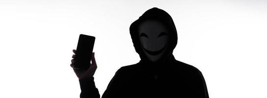 hacker anonym och ansikte mask med smartphone i hand. man i svart huva skjorta innehav och använder sig av mobil telefon på vit bakgrund. representera cyber brottslighet data dataintrång eller stjäla personlig data begrepp foto