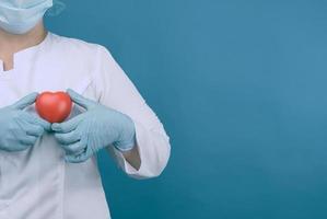 kvinna läkare i en vit täcka, en mask står och innehar en röd hjärta på en blå bakgrund foto