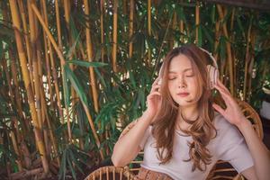 avslappnad asiatisk kvinna med trådlös hörlurar, lyssnande till meditation musik med stängd ögon, leende med nöje. glad ung flicka bär vit t-shirt Sammanträde på stol i mysigt botanisk trädgård. foto