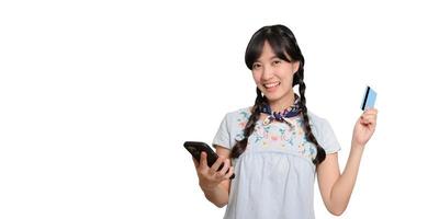 porträtt av skön Lycklig ung asiatisk kvinna i denim klänning innehav kreditera kort och smartphone på vit bakgrund. studio skott foto