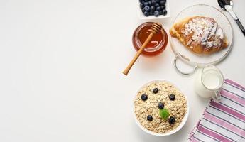 morgon- frukost, rå gröt flingor i en keramisk tallrik, mjölk i en karaff, blåbär och honung i en burk på en vit tabell, topp se foto