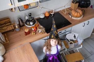 barn dekorerar de kök i Hem för halloween. flicka i en häxa kostym spelar med de dekor för de Semester - fladdermöss, domkraft lykta, pumpor. höst bekvämlighet i hus, scandi-stil kök, loft foto