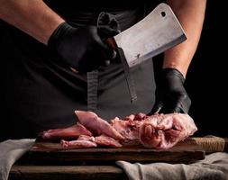 kock i svart latex handskar innehar en stor kniv och nedskärningar in i bitar rå kanin kött på en brun trä- skärande styrelse foto