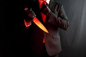 affärsman i mörk kostym och röd slips innehav skarp kniv på svart bakgrund. halloween Skräck film mördare. foto
