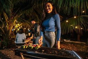porträtt av Lycklig asiatisk ung kvinna matlagning kött på utegrill grill på ny år fest. bar-bq eller bbq på traditionell spis. natt fest, människor och firande begrepp. foto