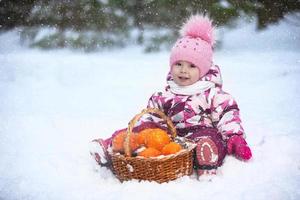 en liten flicka i vinter- kläder är Sammanträde på de snö med en korg av apelsiner. foto