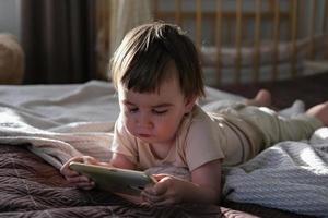 en tvååring pojke lögner på en säng och klockor tecknade serier på en smartphone foto