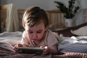 en tvååring pojke lögner på en säng och klockor tecknade serier på en smartphone foto