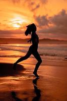 en ballerina med en silhuett form utför balett rörelser mycket flexibelt på de strand med de vågor kraschar foto