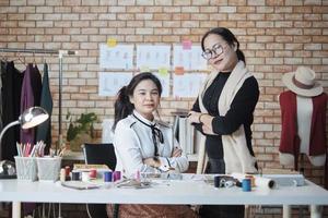 mode team, asiatisk kvinna designer och tonåring assistent i studio, vapen korsade och leende, Lycklig arbetssätt med färgrik tråd och sömnad för klänning design, professionell boutique skräddare sme entreprenör. foto