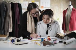 asiatisk medelålders kvinna mode designer lär en ung tonåring praktikant skräddare i studio med färgrik tråd och sömnad tyg för klänning design samling idéer, professionell boutique små företag. foto