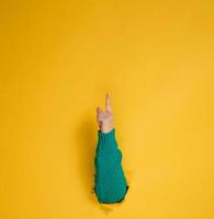 kvinna hand pinnar ut av en trasig hål i en gul papper bakgrund, del av de kropp poäng med de index finger till topp. kopia Plats foto