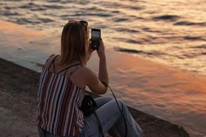 flicka turist fotografier på de telefon en skön vatten landskap på solnedgång foto