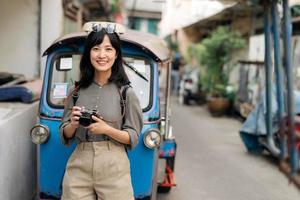 ung asiatisk kvinna ryggsäck resande stående en sida av tuk tuk taxi på sommar semester på Bangkok, thailand. foto