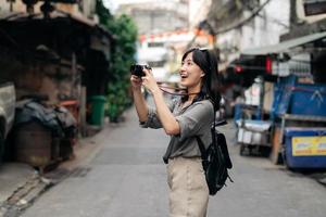 ung asiatisk kvinna ryggsäck resande använder sig av digital kompakt kamera, njuter gata kulturell lokal- plats och leende. foto