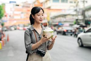 Lycklig ung asiatisk kvinna ryggsäck resande dricka en kokos juice på Kina stad gata mat marknadsföra i Bangkok, thailand. resande kontroll ut sida gator. foto
