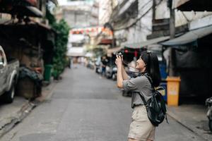 ung asiatisk kvinna ryggsäck resande använder sig av digital kompakt kamera, njuter gata kulturell lokal- plats och leende. foto