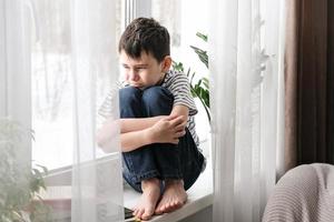 en ledsen pojke sitter på de fönsterkarm kramas hans knäna. dålig humör, depression. de pojke är ledsen ensam på Hem foto