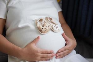 Foto av Lycklig asiatisk gravid kvinna Sammanträde i säng och rörande henne mage på Hem med små skor för de ofödd bebis i de mage