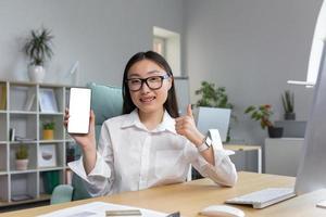 ung asiatisk kvinna i glasögon innehav en mobil telefon i henne hand. som visar de telefon, telefon mockup. foto