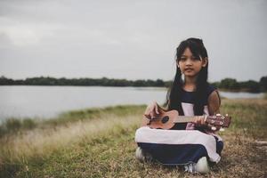 söt liten asiatisk tjej som sitter på gräset och spelar ukulele i parken