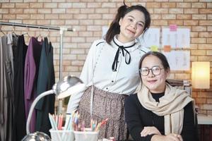 asiatisk kvinna mode designer och ung lärling se på kamera och leende i studio, vapen korsade, arbetssätt med färgrik tråd för klänning design, boutique skräddare sme företag entreprenör. foto