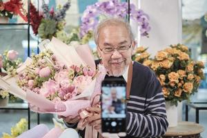 e-handel företag, ett senior manlig blomsterhandlare arbetare demonstrera och visa blommig arrangemang via uppkopplad leva strömning med smartphone Ansökan i en ljus blomma affär, en skön blommar Lagra. foto