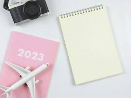 platt lägga av tom sida öppnad anteckningsbok, rosa 2023 dagbok, flygplan modell och kamera på vit bakgrund med kopia Plats. resa, Foto och minne begrepp.