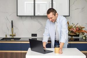 en ung man stående i de kök är arbetssätt på en bärbar dator och innehav en disponibel kopp foto