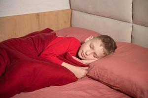 en söt pojke i röd pyjamas är liggande på en kudde med hans ögon stängd i säng och leende foto