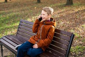 en pojke är talande på de telefon och skrattande medan Sammanträde på en parkera bänk i höst foto
